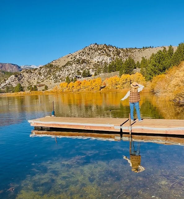 Traveler standing on a wooden dock overlooking Gull Lake in June Lake, California, surrounded by golden fall foliage and mountain scenery under a clear blue sky