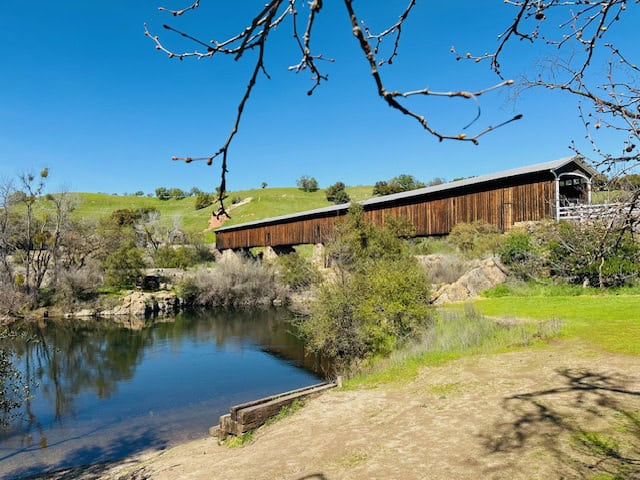 Historic Knights Ferry Covered Bridge spanning the Stanislaus River in California on a sunny spring day, surrounded by green rolling hills and calm reflective water