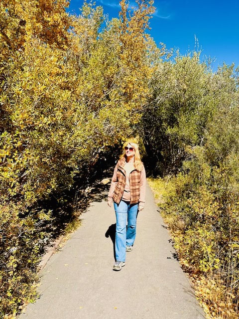 Woman walking along the lakeside path at Convict Lake in California’s Eastern Sierra, surrounded by fall foliage and mountain peaks under a clear blue sky.