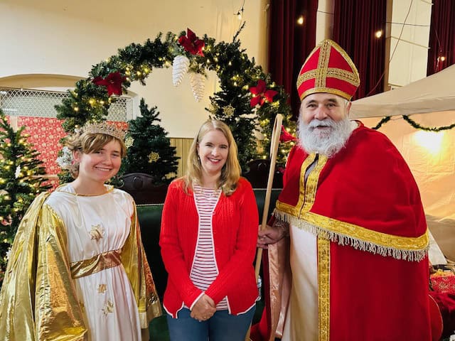Festive scene at the Sacramento Christkindlmarkt with a visitor standing beside St. Nikolaus and the Christkind in front of a holiday wreath and decorated Christmas trees