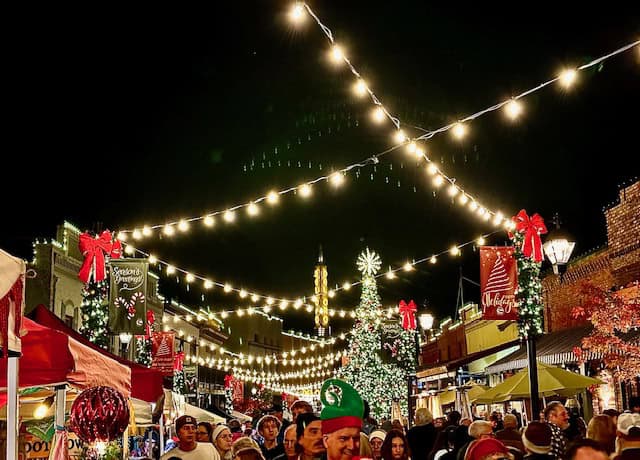 Festive street scene at Cornish Christmas in Grass Valley with string lights, decorated Christmas trees, red bows, vendor booths, and a lively crowd enjoying the holiday market at night.
