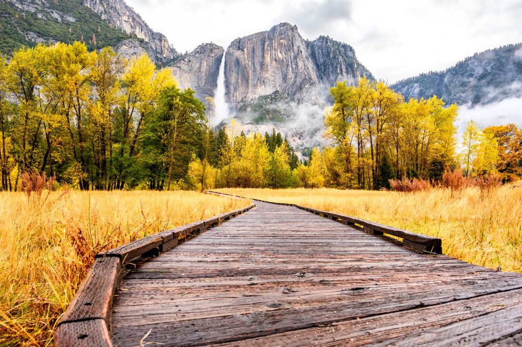 Wooden boardwalk winding through Yosemite Valley with golden fall foliage and Upper Yosemite Fall flowing down the granite cliffs in the background.