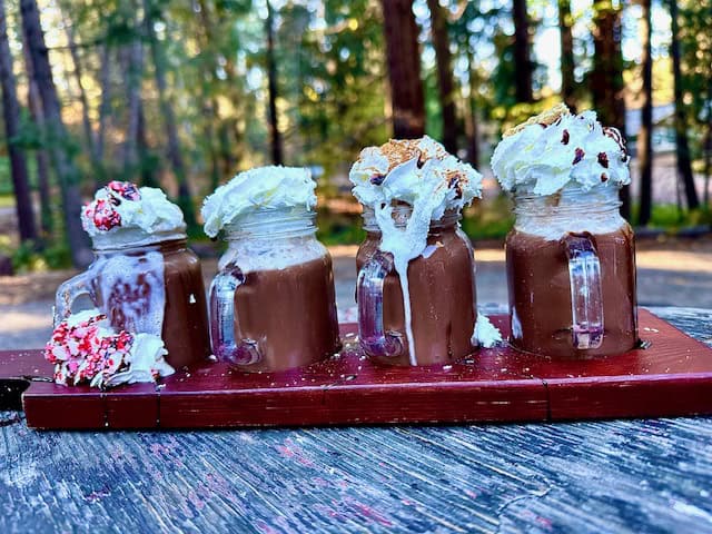 Mini hot chocolate flight topped with whipped cream and festive toppings at Grandpa’s Cellar in Apple Hill, displayed outdoors on a rustic wooden table.