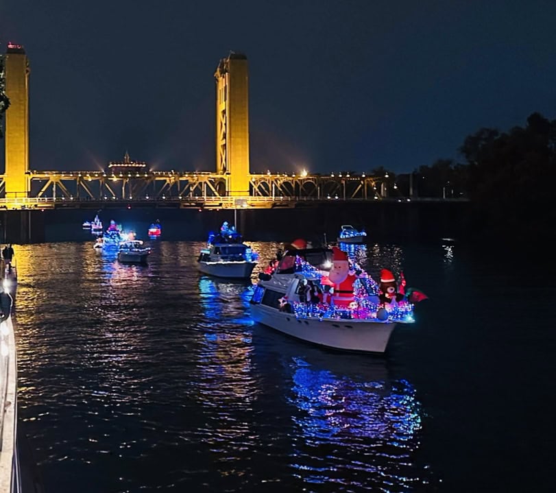 Holiday boat parade on the Sacramento River with colorful Christmas lights sailing past the illuminated Tower Bridge, viewed from a festive river cruise