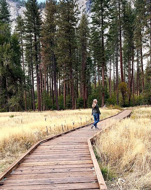 Woman walking along a wooden boardwalk through a golden meadow in Yosemite Valley with tall pines and granite cliffs in the background.