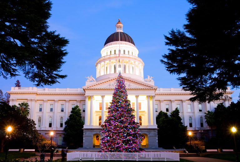 A beautifully lit Christmas tree in front of the California State Capitol in Sacramento at dusk, with holiday lights glowing against the historic white building.