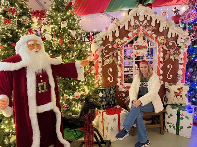Woman sitting beside a festive gingerbread house and Christmas trees at the Cambria Christmas Market in Cambria, California, with a Santa statue nearby