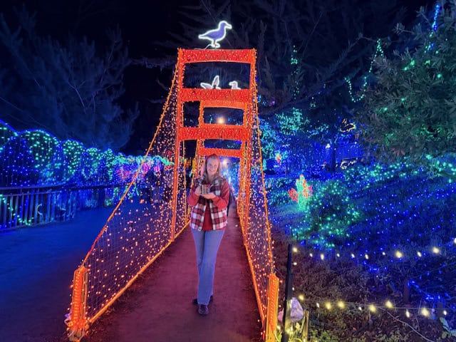 Woman walking across a festive lighted bridge holding a collectible mug filled with hot cocoa at the Cambria Christmas Market in California, surrounded by colorful holiday lights at night