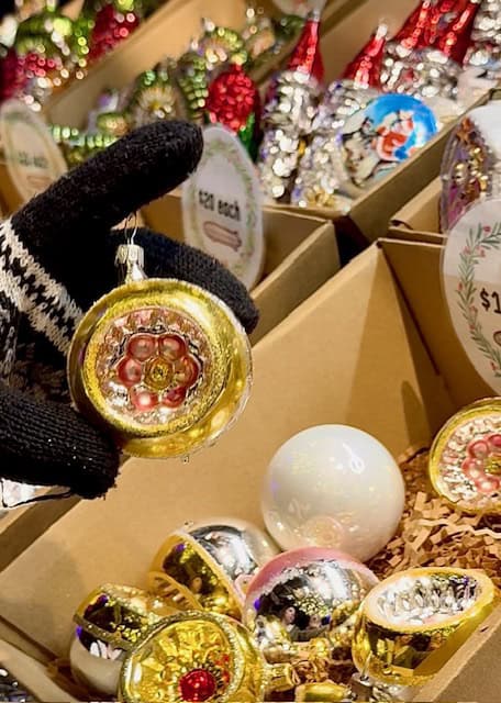 Hand holding a hand-blown glass Christmas ornament from Germany at the Cambria Christmas Market, surrounded by colorful traditional holiday ornaments