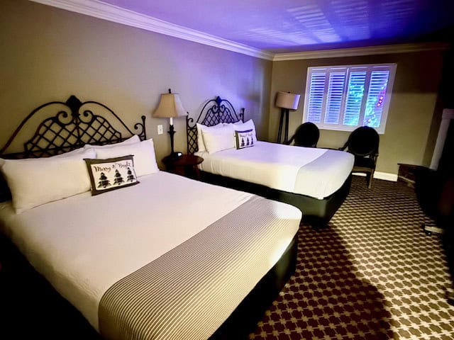 Two queen beds in a cozy guest room at Cambria Pines Lodge, featuring wrought-iron headboards, neutral décor, and warm lighting