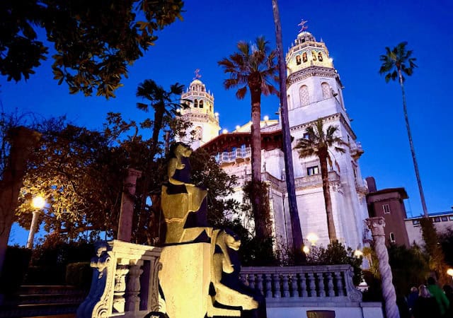 Night view of Hearst Castle decorated for Christmas, with illuminated towers, palm trees, and holiday lights against a deep blue evening sky