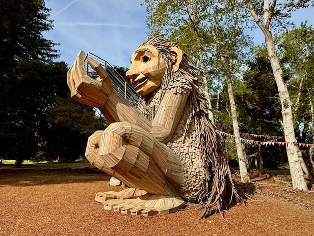 Large wooden troll sculpture at Filoli Gardens in California, handcrafted from recycled wood with detailed face and seated pose among trees