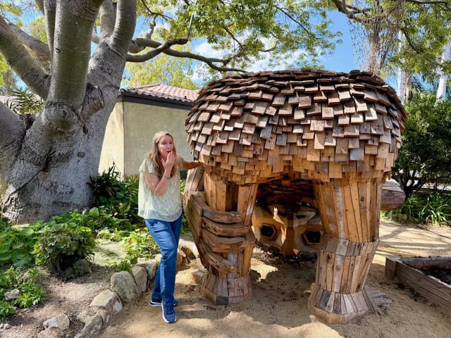 Woman posing with upside-down wooden troll sculpture made of recycled wood in Solvang California, whimsical outdoor art installation under trees