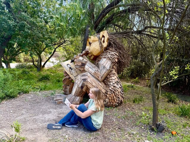 Woman sitting near wooden troll sculpture reading a map at South Coast Botanic Gardens, large recycled wood art figure drawing a picture in forest setting