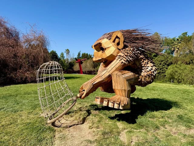 Wooden troll sculpture using a net in an open grassy field at South Coast Botanic Gardens, large recycled wood art surrounded by greenery