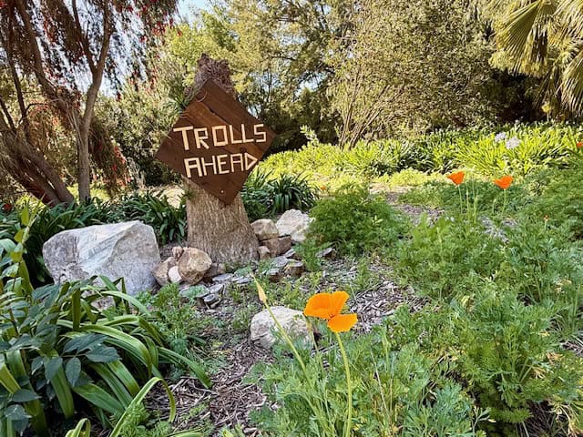 Whimsical “Trolls Ahead” wooden sign along a garden path with blooming orange California poppies and lush greenery in a California park