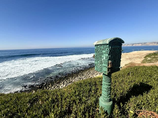 Scenic ocean view along the La Jolla coastline featuring waves crashing against rocky shores, coastal cliffs, and a historic green lifeguard call box under a clear blue sky