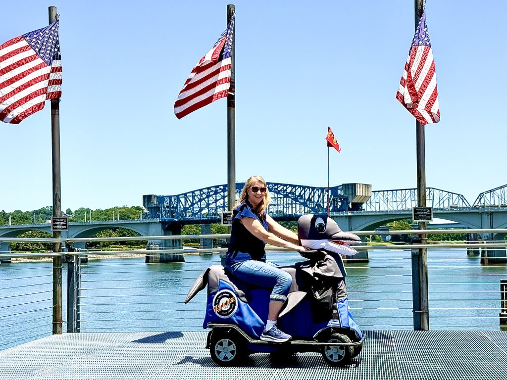 Traveler riding a Rydables shark scooter along the Chattanooga Riverwalk with the Walnut Street Bridge and Tennessee River in the background