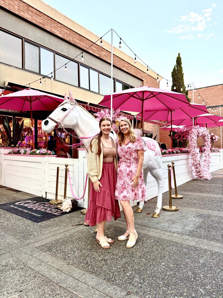 Two women outside Café Carrusel in Santa Rosa with a white carousel horse, bright pink umbrellas, and floral decorations